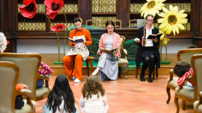 Lettura animata e laboratorio creativo per bambini con scenografia floreale (fiori giganti, papaveri rossi e margherite gialle) sullo sfondo e bambini seduti